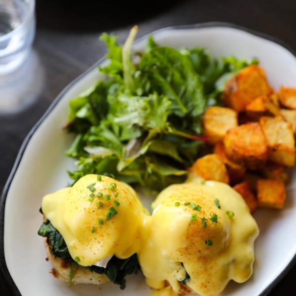 Two eggs Benedict with hollandaise sauce, chives, a side salad, and roasted potatoes.