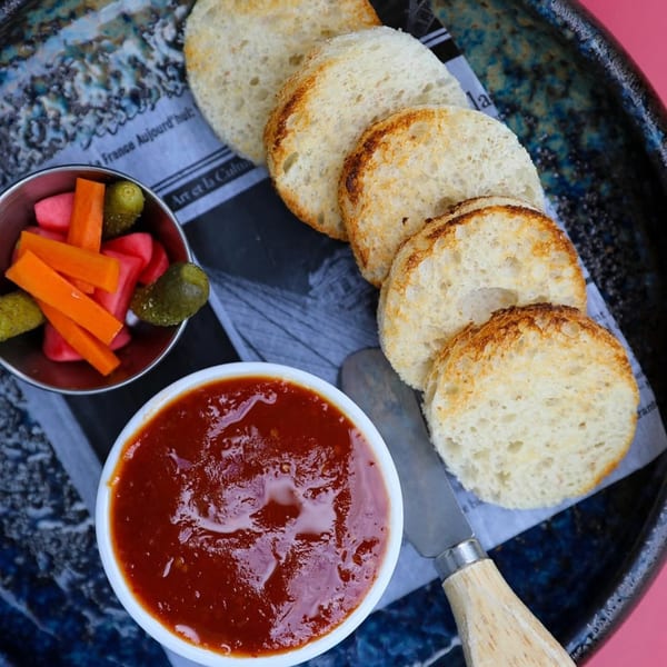 Toasted baguette slices, red sauce, and pickled vegetables on a dark blue plate with newspaper.